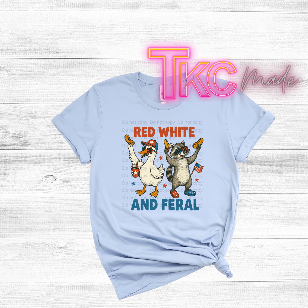 Light blue t-shirt with a graphic of a duck and raccoon holding American flags and text 'Red White and Feral' on a wooden surface.
