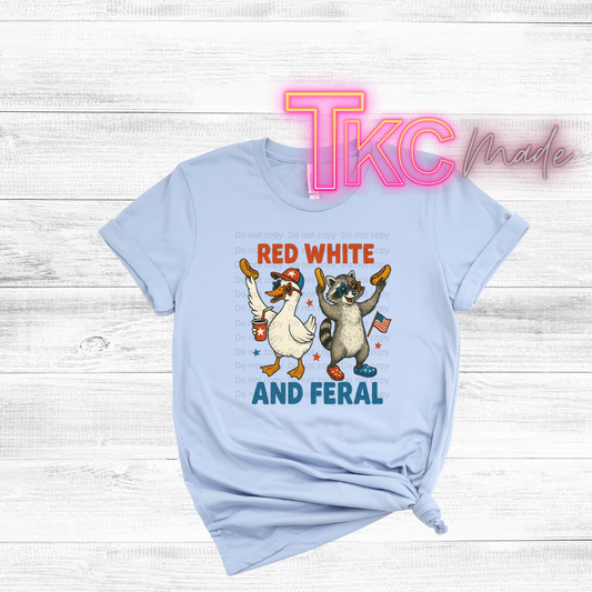 Light blue t-shirt with a graphic of a duck and raccoon holding American flags and text 'Red White and Feral' on a wooden surface.
