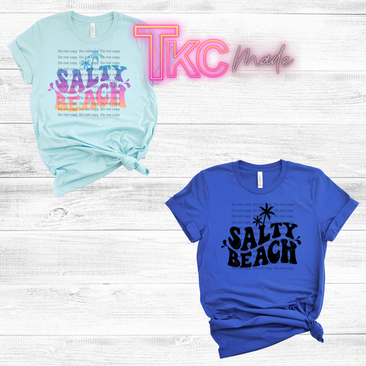 Two t-shirts with 'Salty Beach' text on a wooden surface, featuring the brand 'TKC Made'.