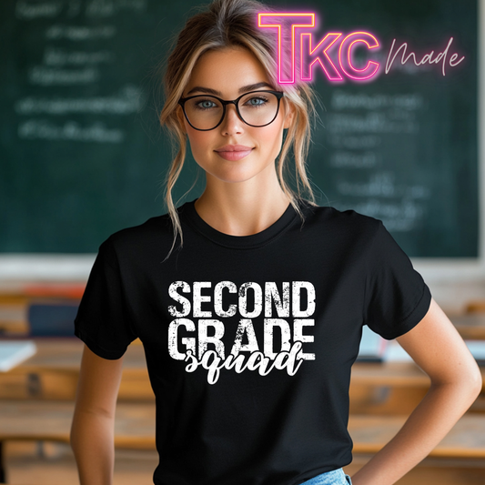 A woman wearing a black graphic t-shirt with the text 'SECOND GRADE squad' in white, standing in front of a chalkboard. A pink and yellow 'TKC made' watermark is visible in the top right corner.