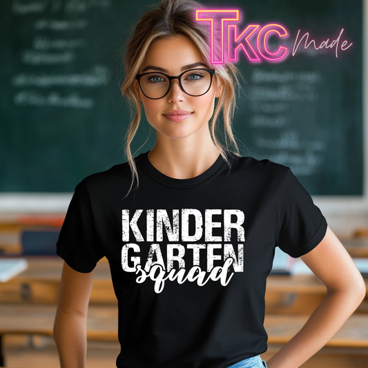 A woman wearing a black graphic t-shirt with the text 'KINDERGARTEN squad' in white, standing in front of a blurred chalkboard background. A pink and yellow 'TKC made' watermark is visible in the top right corner.