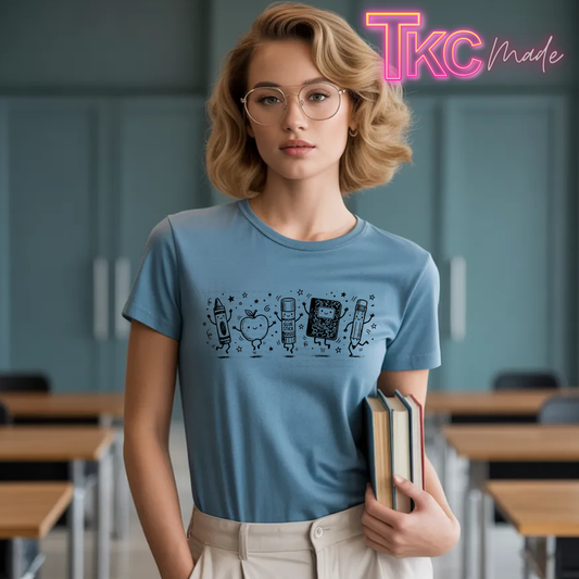 A woman with blonde wavy hair and glasses stands in a classroom holding books, wearing a light blue crewneck t-shirt with a black school-themed graphic print. The background shows blurred desks and cabinets.