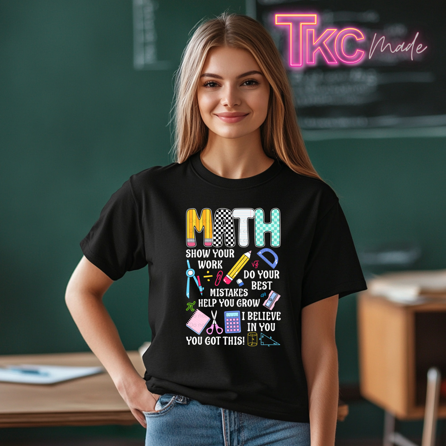 A woman wearing a black graphic t-shirt with a colorful 'MATH' design and motivational text, standing in a classroom setting with a chalkboard in the background.