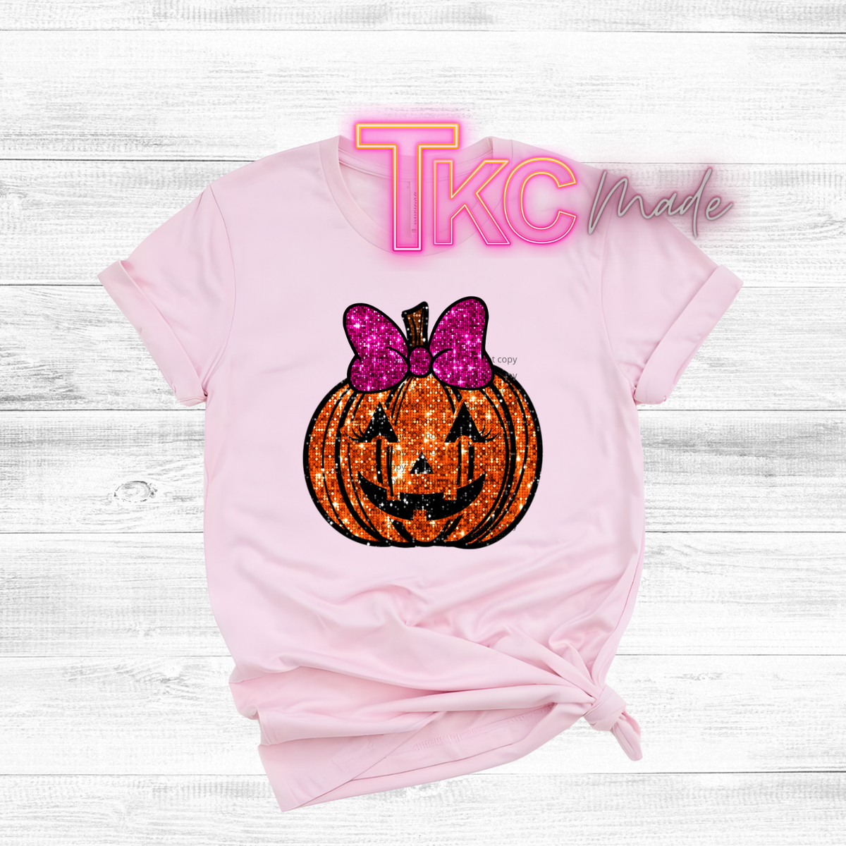A light pink short-sleeve t-shirt with a Halloween graphic of a glittery orange pumpkin jack-o'-lantern wearing a pink glitter bow. The shirt is styled with a knot at the bottom hem and is laid flat on a white wood plank background.
