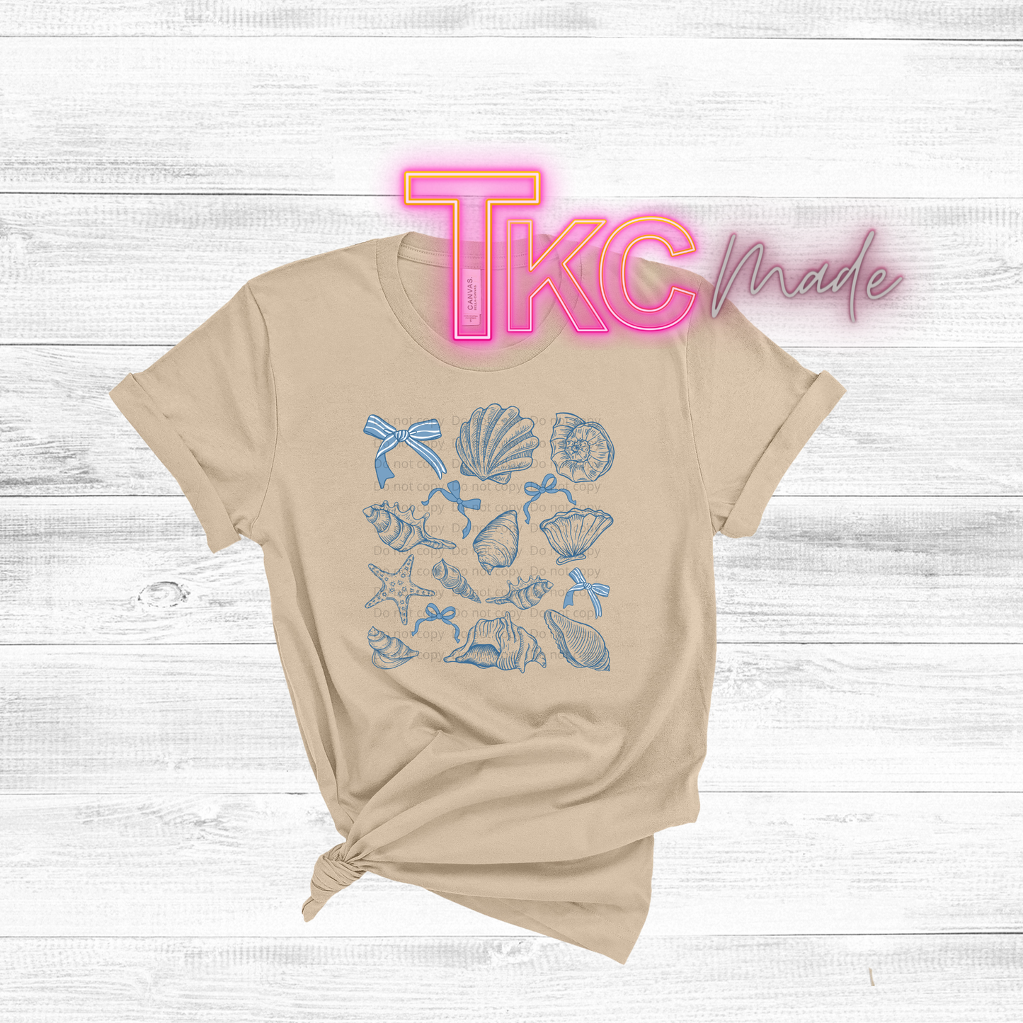 Beige t-shirt with blue shell and bow design on a white wooden background with 'TKC Made' text.
