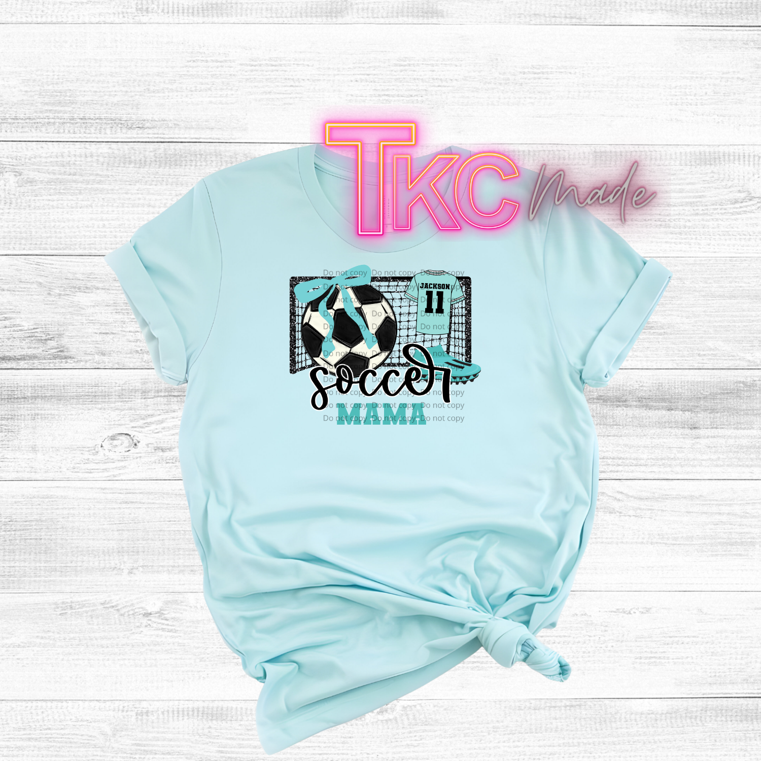 Light blue t-shirt with 'Soccer Mama' design  with a goalie and 'TKC made' text on a white wooden background