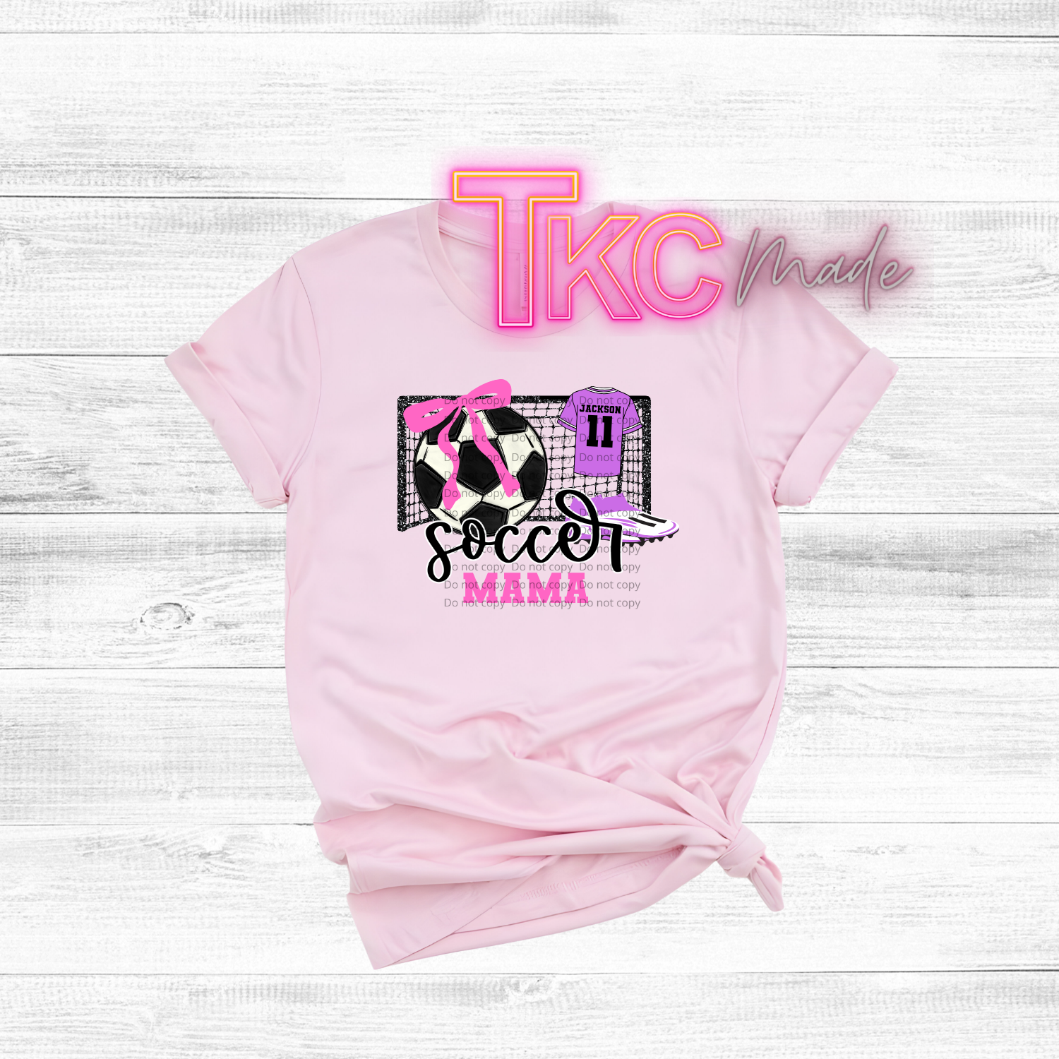 Pink t-shirt with 'Soccer Mama' design and 'TKC made' text on a white wooden background
