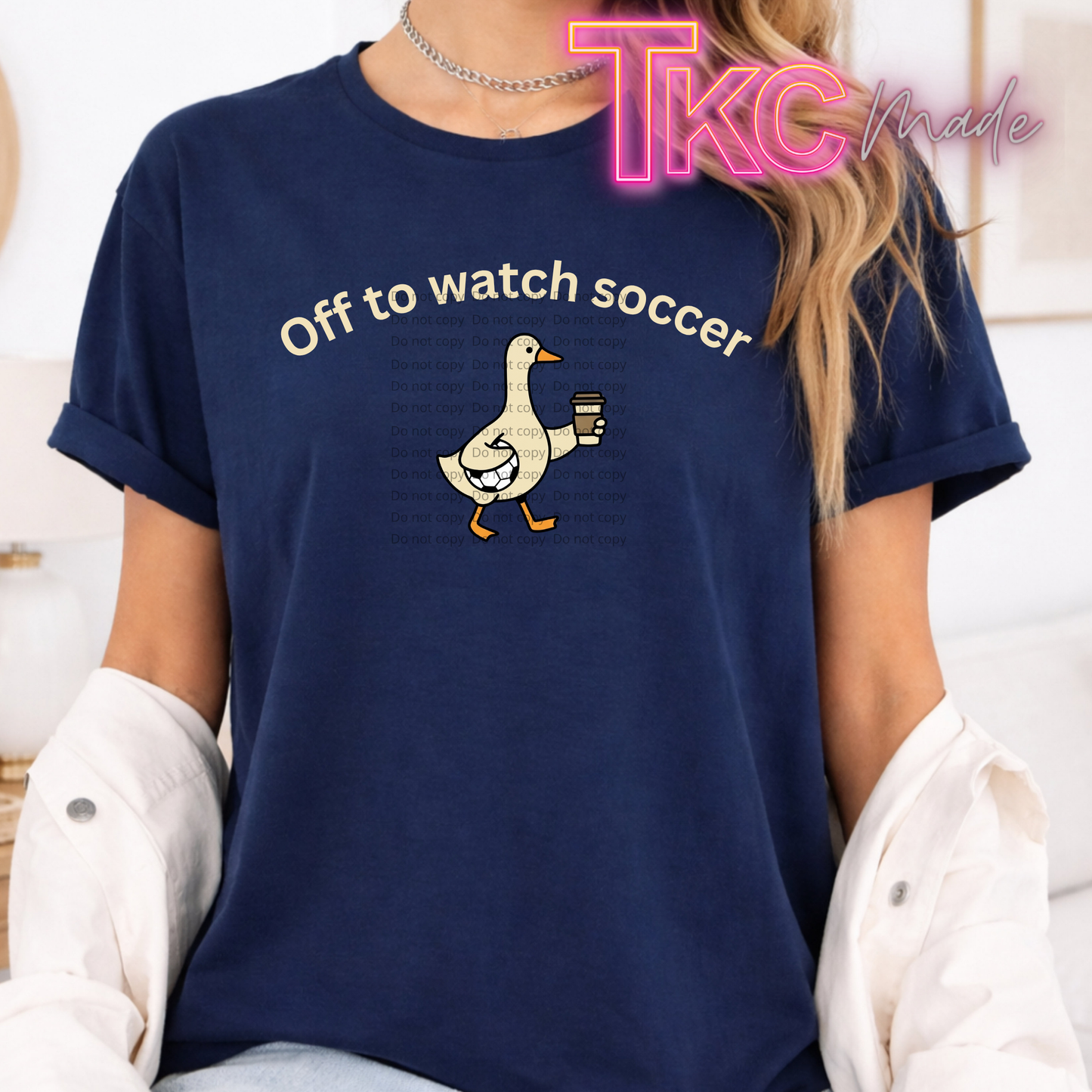 A woman wearing a navy blue graphic t-shirt with the text 'Off to watch soccer' and a cartoon duck holding a coffee cup. A white button-down shirt is casually draped off her shoulders, and a 'TKC Made' watermark is visible in the top right corner.
