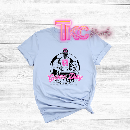 Light blue t-shirt with sports-themed soccer girl design and 'TKC made' text on a white wooden background