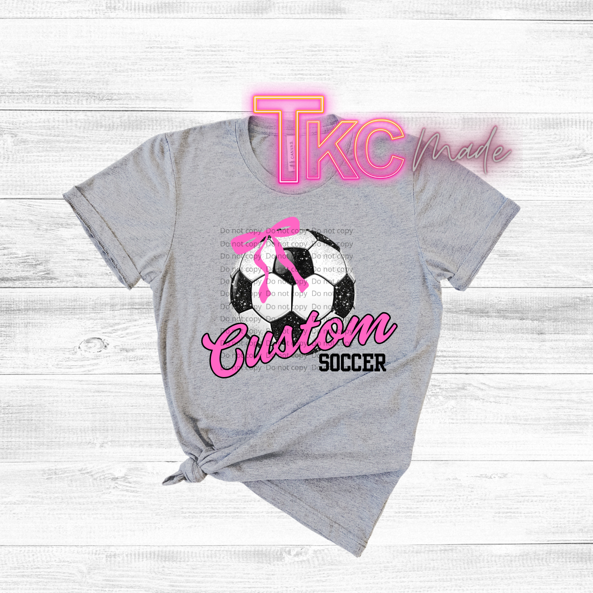 Gray t-shirt with soccer ball and pink bow design, featuring 'TKC made' text on a light wooden background.