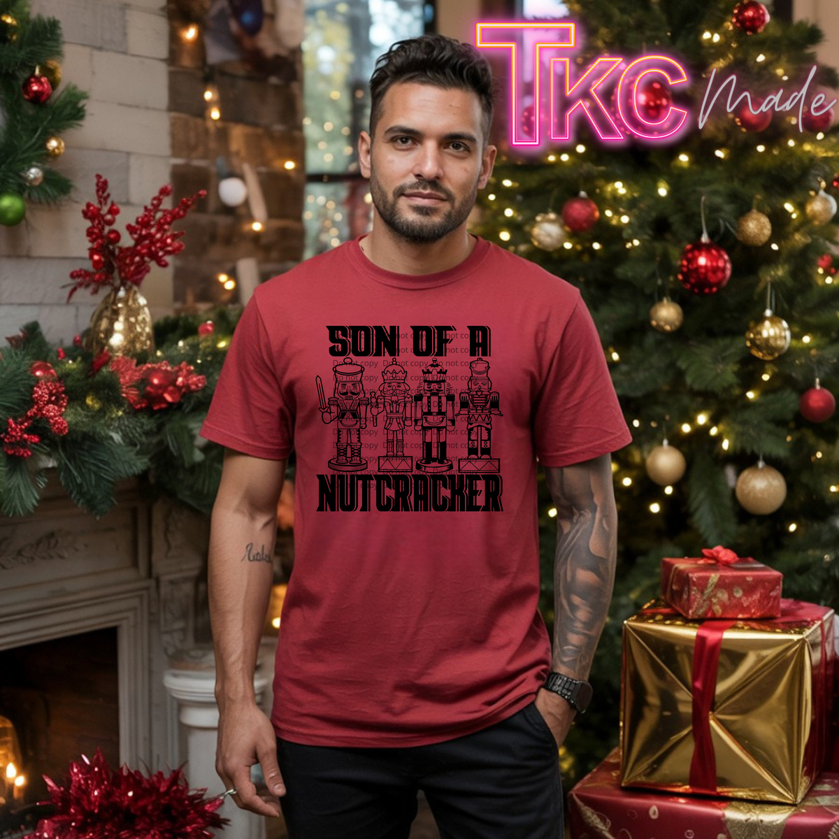 A man wearing a red graphic t-shirt with a black 'SON OF A NUTCRACHER' design, standing in a decorated Christmas setting with a fireplace, tree, and wrapped gifts. A 'Tkc Made' watermark is visible in the top right corner.