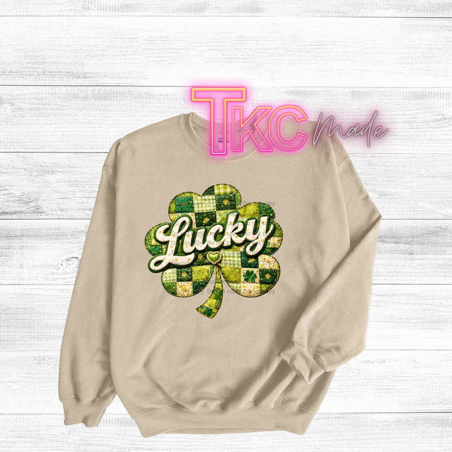 Beige sweatshirt with a green and yellow shamrock design and 'Lucky' text on a white wooden background.