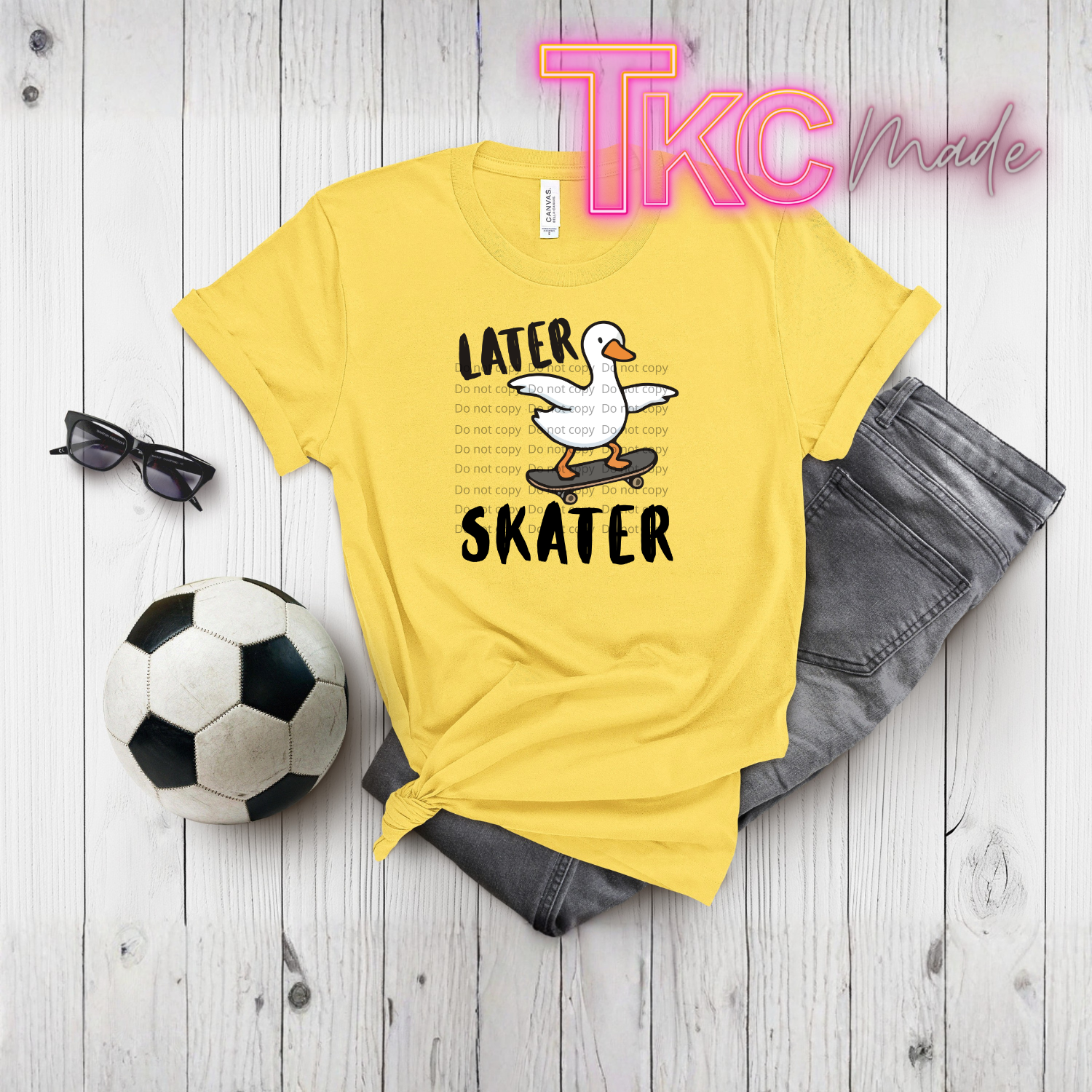 Yellow t-shirt with a graphic and text later skater with a duck, sunglasses, soccer ball, and jeans on a wooden surface with 'TKC Made' branding.