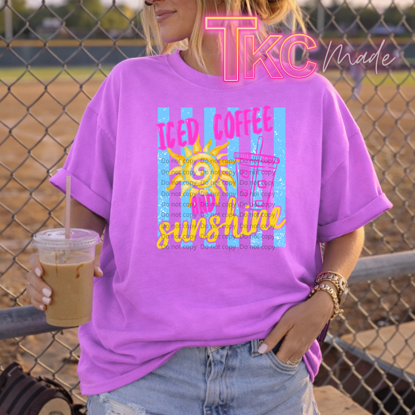 Person wearing a purple t-shirt with a graphic design and holding a drink, standing in front of a chain-link fence.