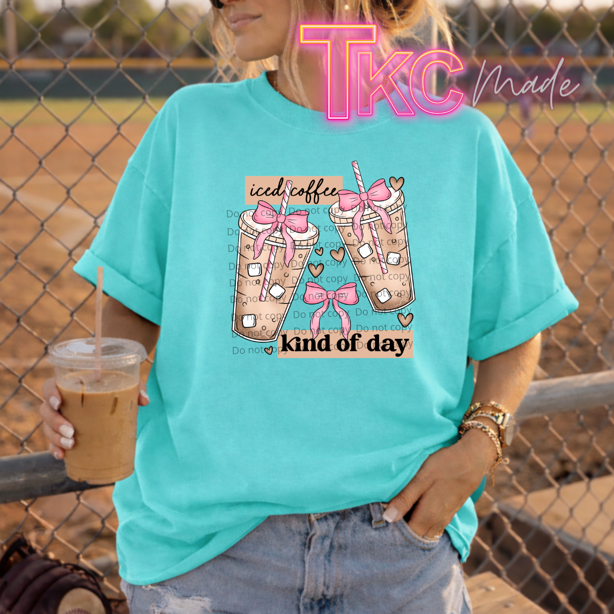 Person wearing a teal t-shirt with a graphic design and holding an iced coffee, standing in front of a chain-link fence.