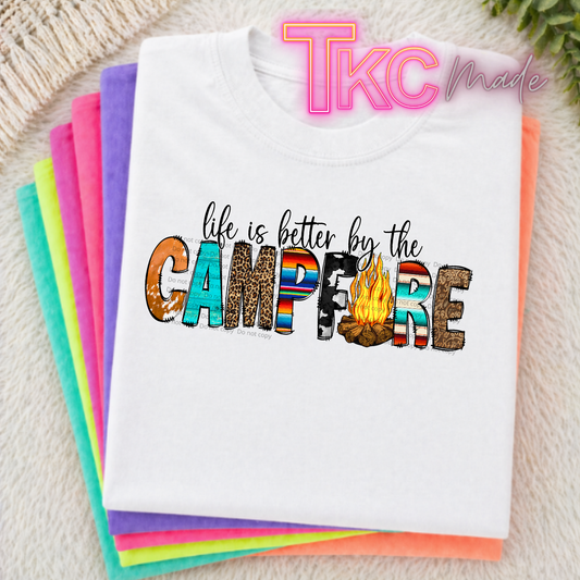 Stack of colorful t-shirts with 'life is better by the campfire' text on a textured surface.