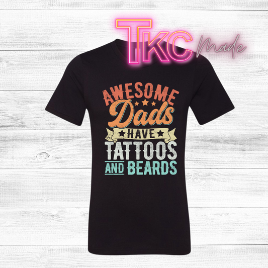 Black t-shirt with colorful text  that reads awesome dads have tattoos and beards on a white wooden background