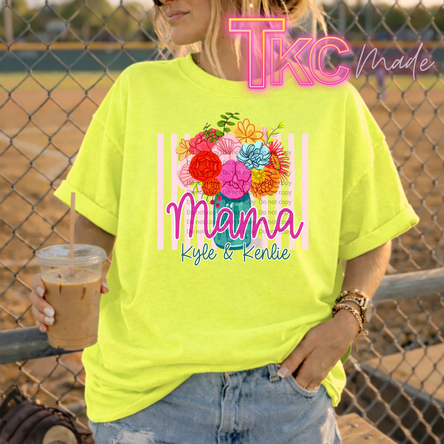 Person wearing a bright yellow t-shirt with floral design and text showing mama and kids names, holding a drink, standing in front of a chain-link fence.