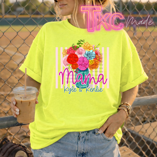 Person wearing a bright yellow t-shirt with floral design and text showing mama and kids names, holding a drink, standing in front of a chain-link fence.