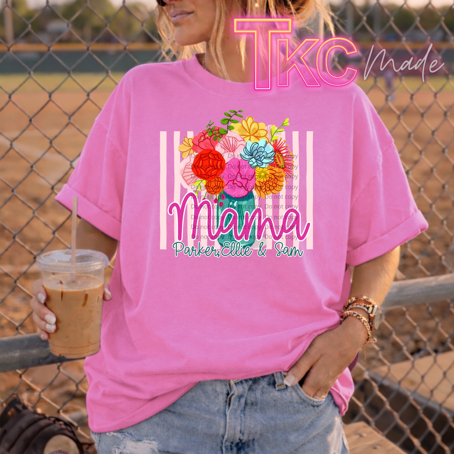 Person wearing a pink t-shirt with floral design and text showing mama and kids names, holding a drink, standing in front of a chain-link fence.