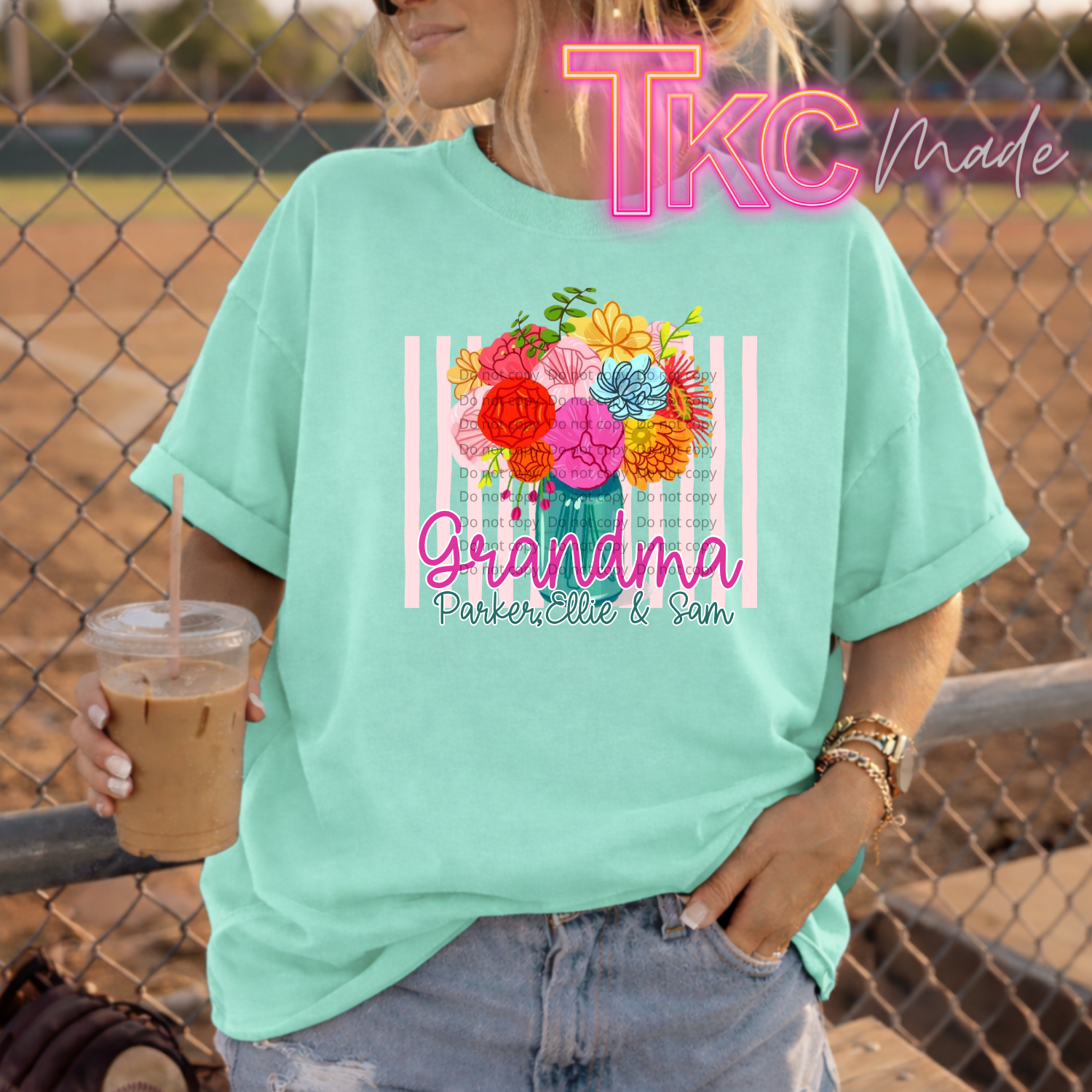 Person wearing a mint t-shirt with floral design and text showing Grandma and kids names, holding a drink, standing in front of a chain-link fence.