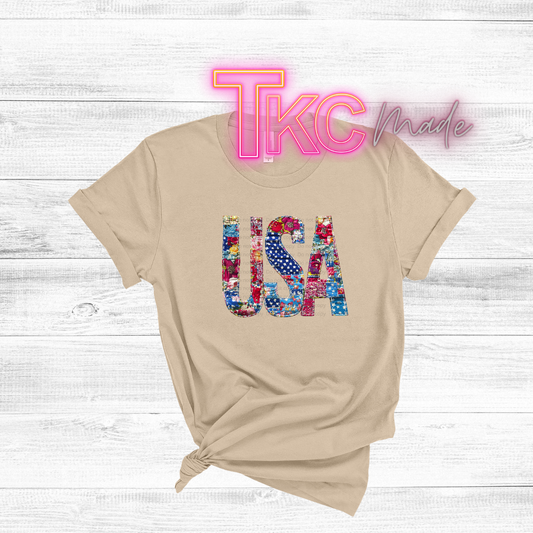 Beige t-shirt with colorful 'USA' design and 'TKC Made' text on a light wooden background