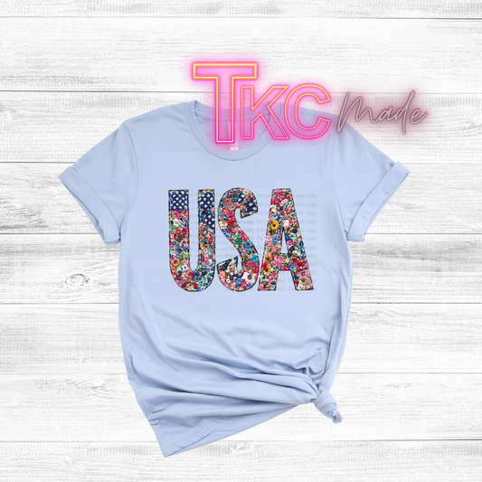 Light blue t-shirt with floral 'USA' design on a wooden surface, featuring 'TKC Made' text.