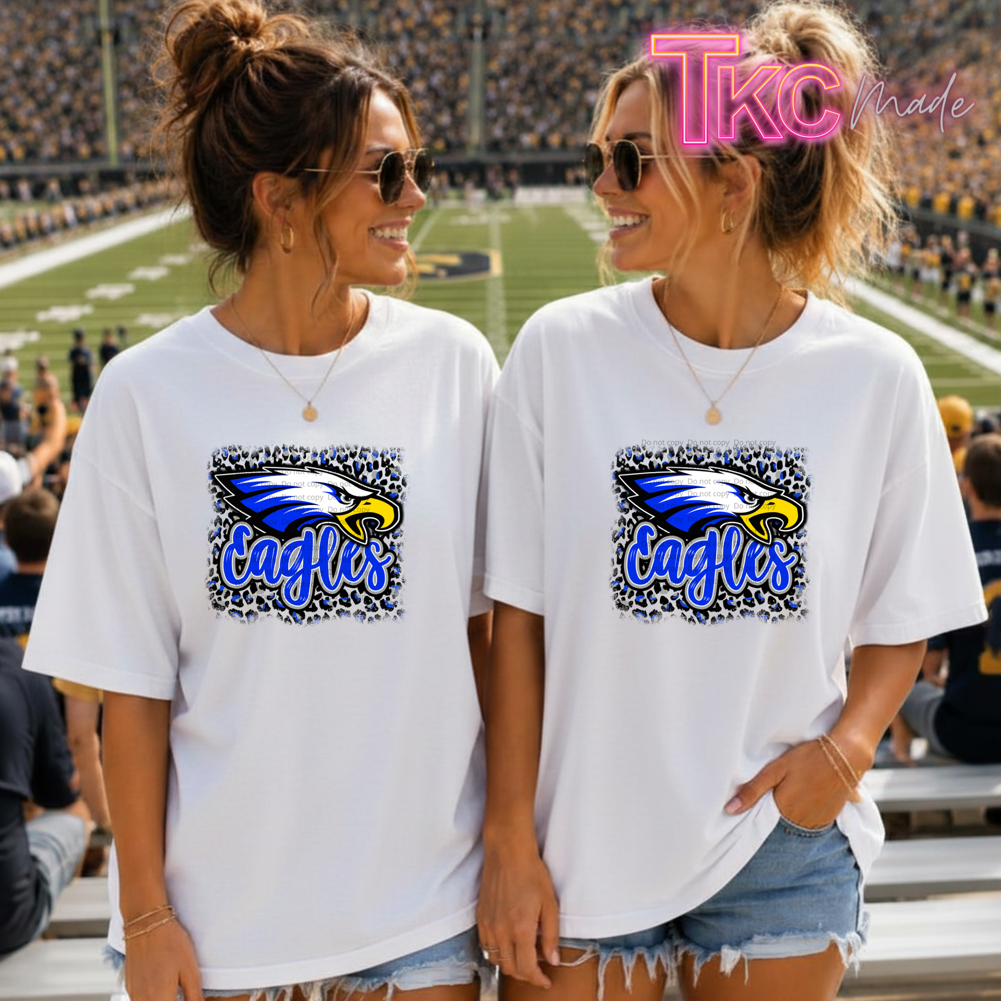 Two women standing together in a stadium wearing matching white graphic t-shirts with a blue and yellow eagles mascot with leopard print