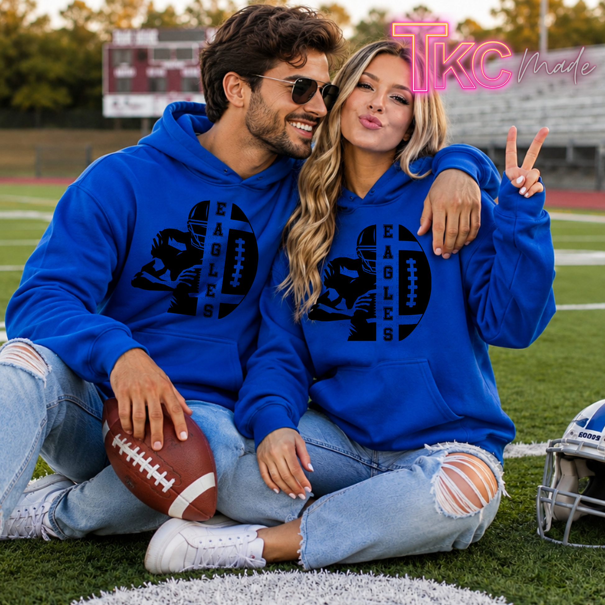 A man and a woman sitting together on a football field wearing matching royal blue hoodies with a black graphic of an eagle head and the word 'EAGLES'. The man holds a football, and a helmet is visible on the right.