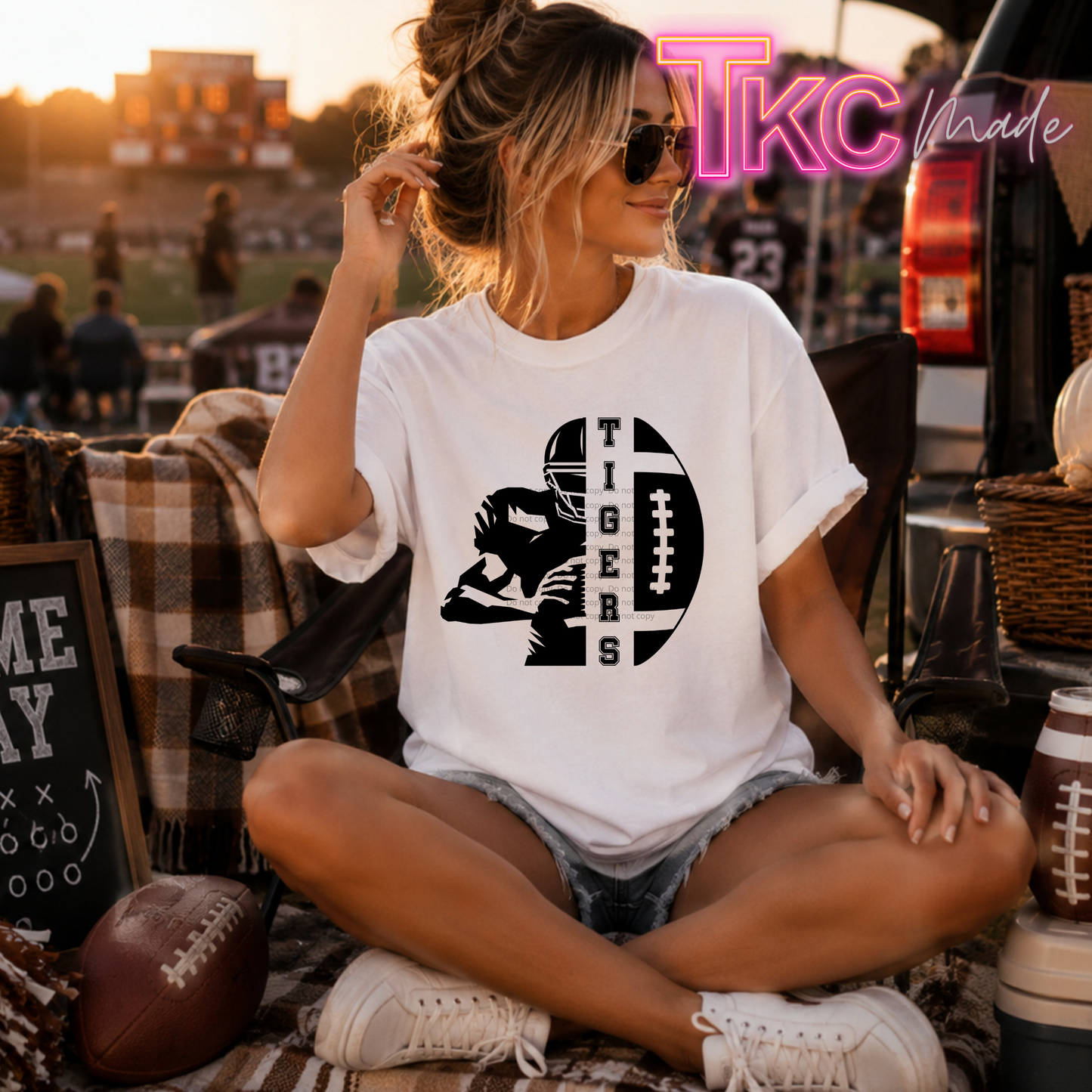 A woman sitting cross-legged outdoors at sunset wearing a white graphic T-shirt with a black football player and the word 'TIGERS' printed on the front. The scene includes a football, plaid blanket, chair, and a 'TKC Made' watermark/logo in the upper right.