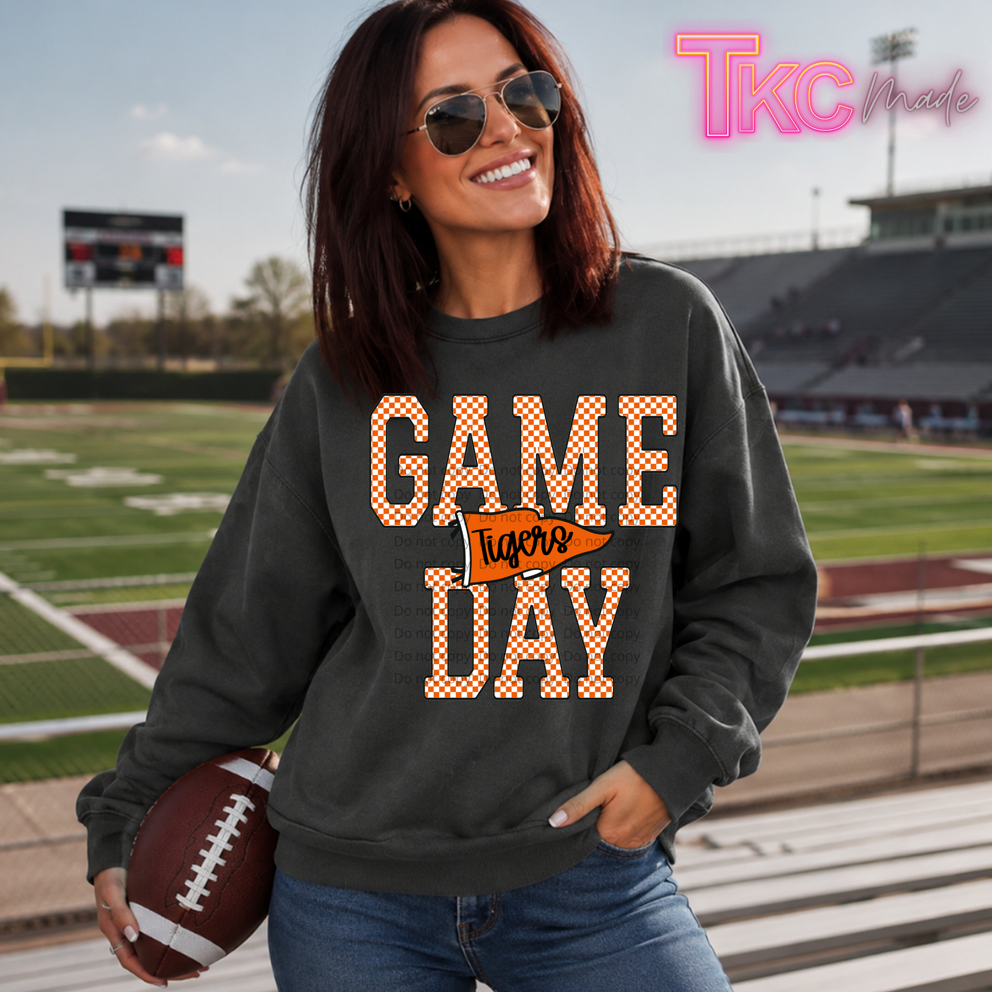 A woman wearing a dark charcoal gray sweatshirt with a 'GAME TIGERS DAY' graphic in orange and white gingham pattern stands on a football field holding a football. The sweatshirt features a small orange arrowhead graphic with 'Tigers' written in script.