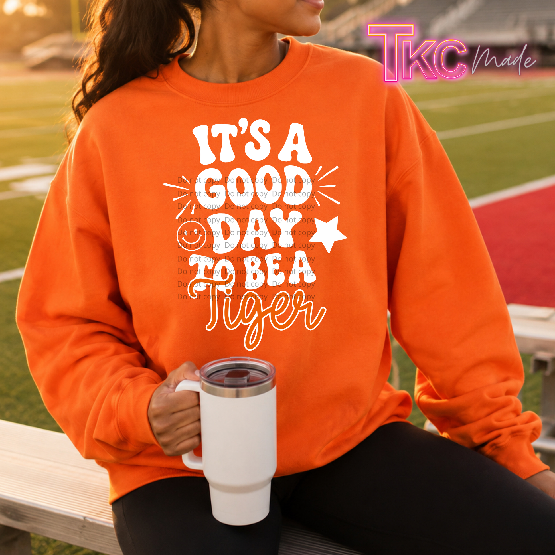 A woman sitting on a bench at a sports field wearing a bright orange sweatshirt with a white graphic that reads 'IT'S A GOOD DAY TO BE A Tiger'. She is holding a white travel mug.