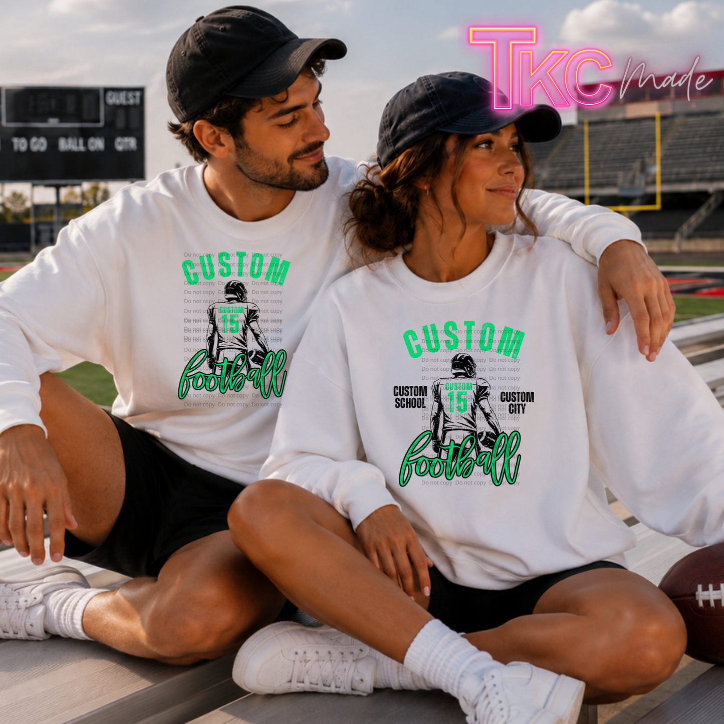 A man and a woman sitting together on a stadium bench, both wearing matching white long-sleeve graphic t-shirts with green and black football-themed custom prints. The background shows a football field and stadium seating under a cloudy sky.