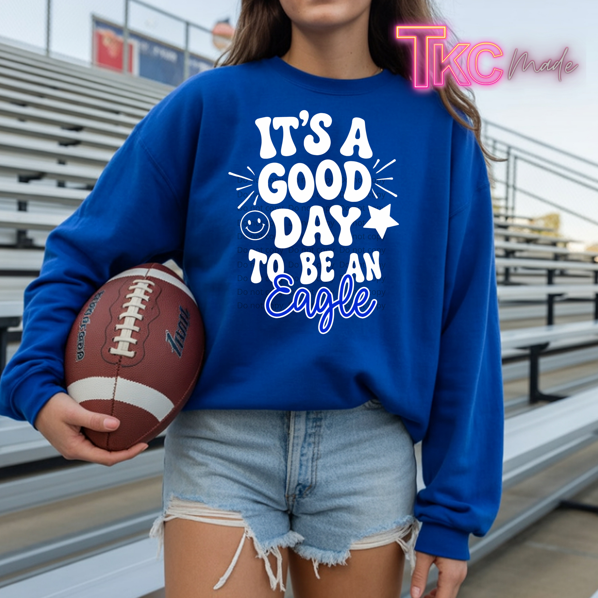 A person wearing a royal blue sweatshirt with a white and blue graphic that reads 'IT'S A GOOD DAY TO BE AN Eagle' stands in a stadium holding a football. The person is also wearing light-wash denim shorts.