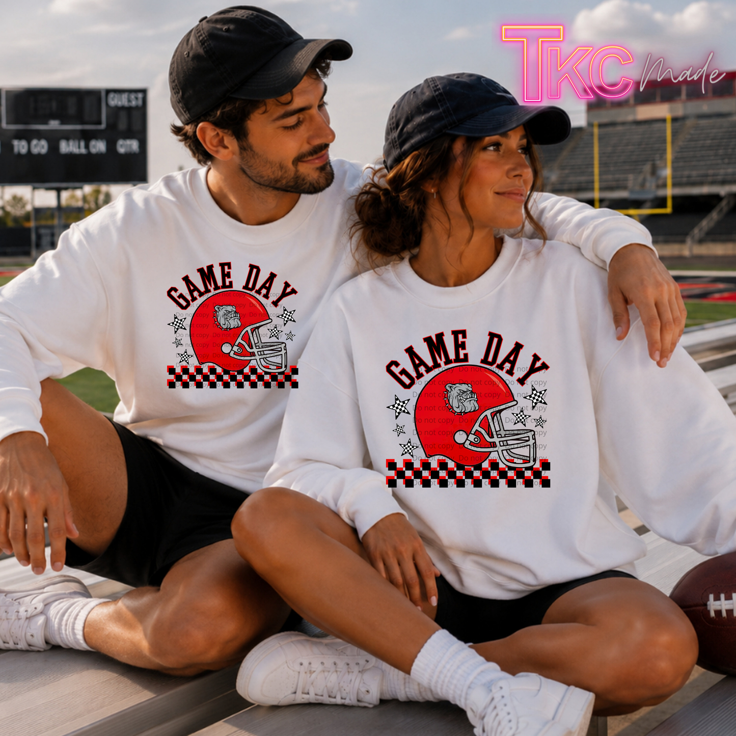A man and a woman sitting together on stadium bleachers wearing matching white long-sleeve graphic shirts with a red football helmet and the words 'GAME DAY' on the front with a bulldogs football. The photo is taken at a football field, with a pink 'TkcMade' watermark in the top right corner.