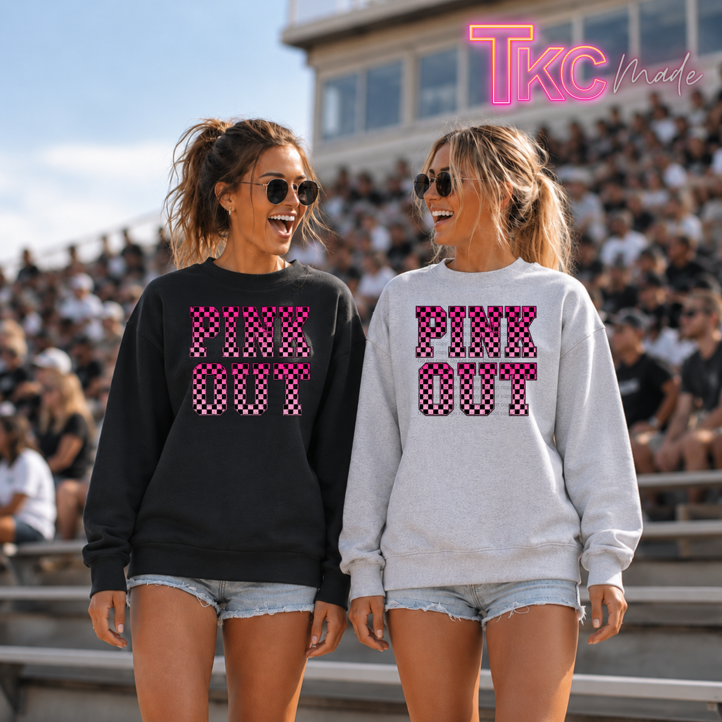 Two women standing in outdoor stadium seating wearing matching sweatshirts with 'PINK OUT' text in a pink-and-white pattern. One sweatshirt is black and the other is light gray.