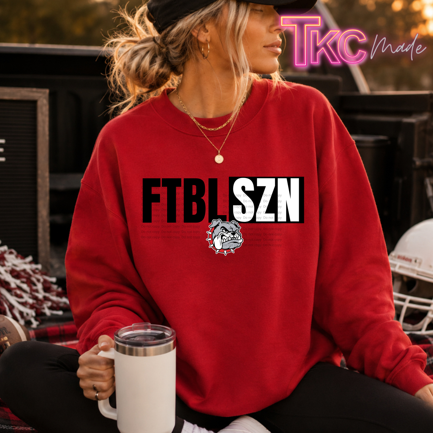 A woman wearing a red crewneck sweatshirt with 'FTBL SZN' and a bulldog graphic printed on the front, sitting outdoors and holding a white mug. A 'Tkc Made' watermark is visible in the top right corner.