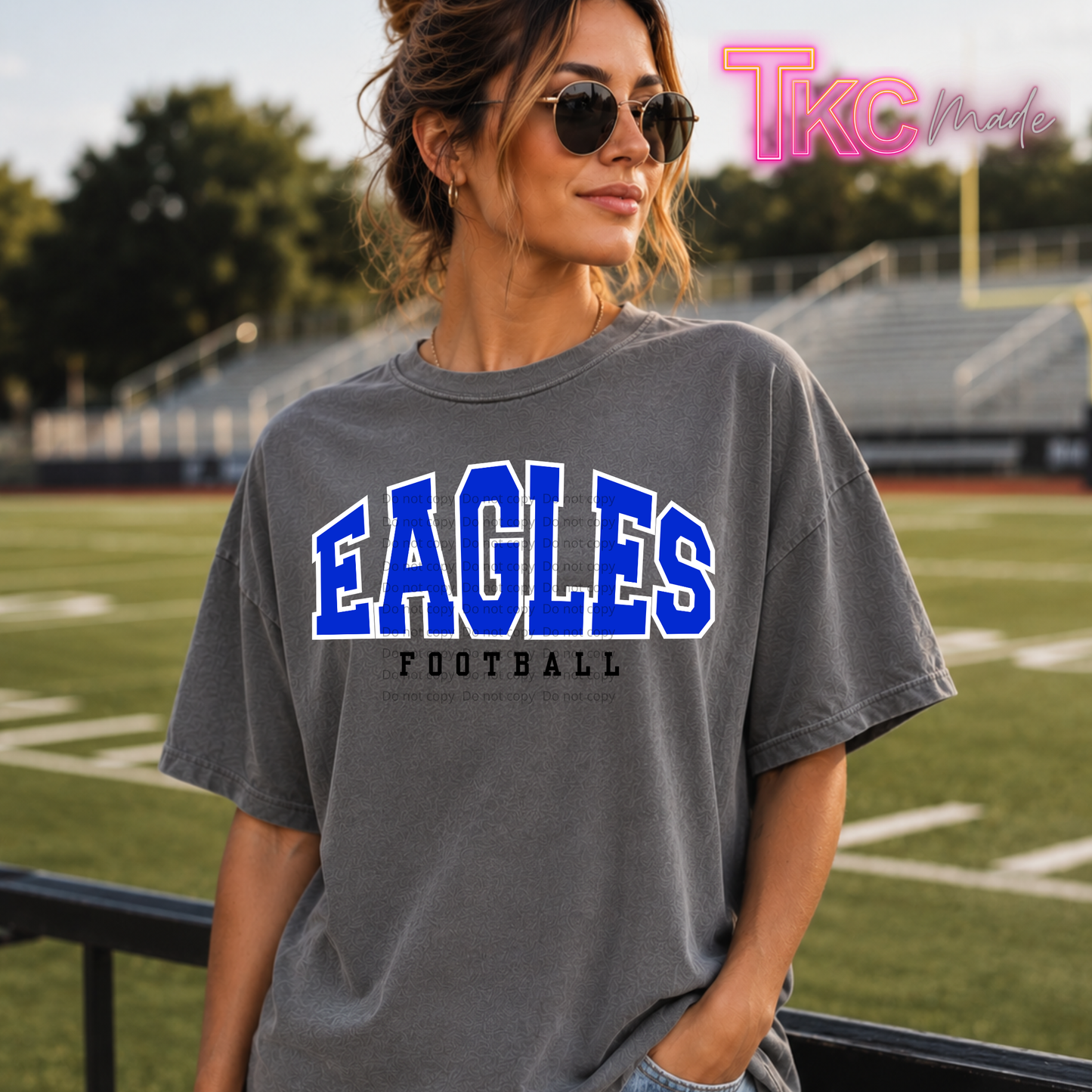 A woman wearing a faded dark gray graphic t-shirt with 'EAGLES FOOTBALL' printed in blue and white on the front, standing on a football field with stadium bleachers in the background.