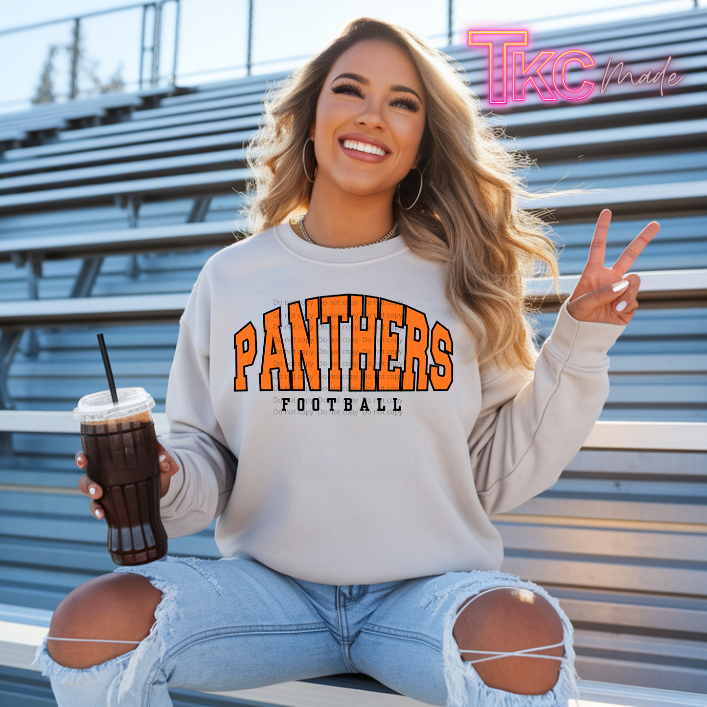 A woman sitting on outdoor stadium bleachers wears a light gray sweatshirt with 'PANTHERS FOOTBALL' printed in orange and black across the chest. She holds an iced coffee and makes a peace sign, with a pink 'TKC Brand' logo in the top right corner.