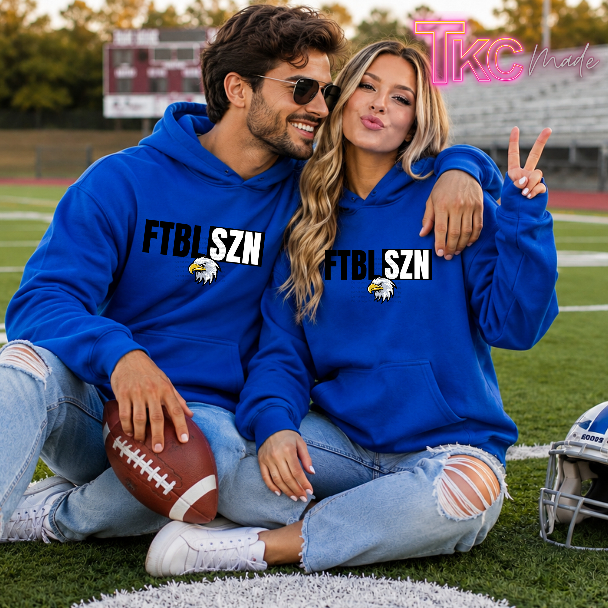 A man and a woman sitting together on a football field wearing matching royal blue hoodies with 'FTBLSZN' and an eagle graphic printed on the front. The man holds a football, and a pink 'TTC Nicole' watermark is visible in the top right corner.