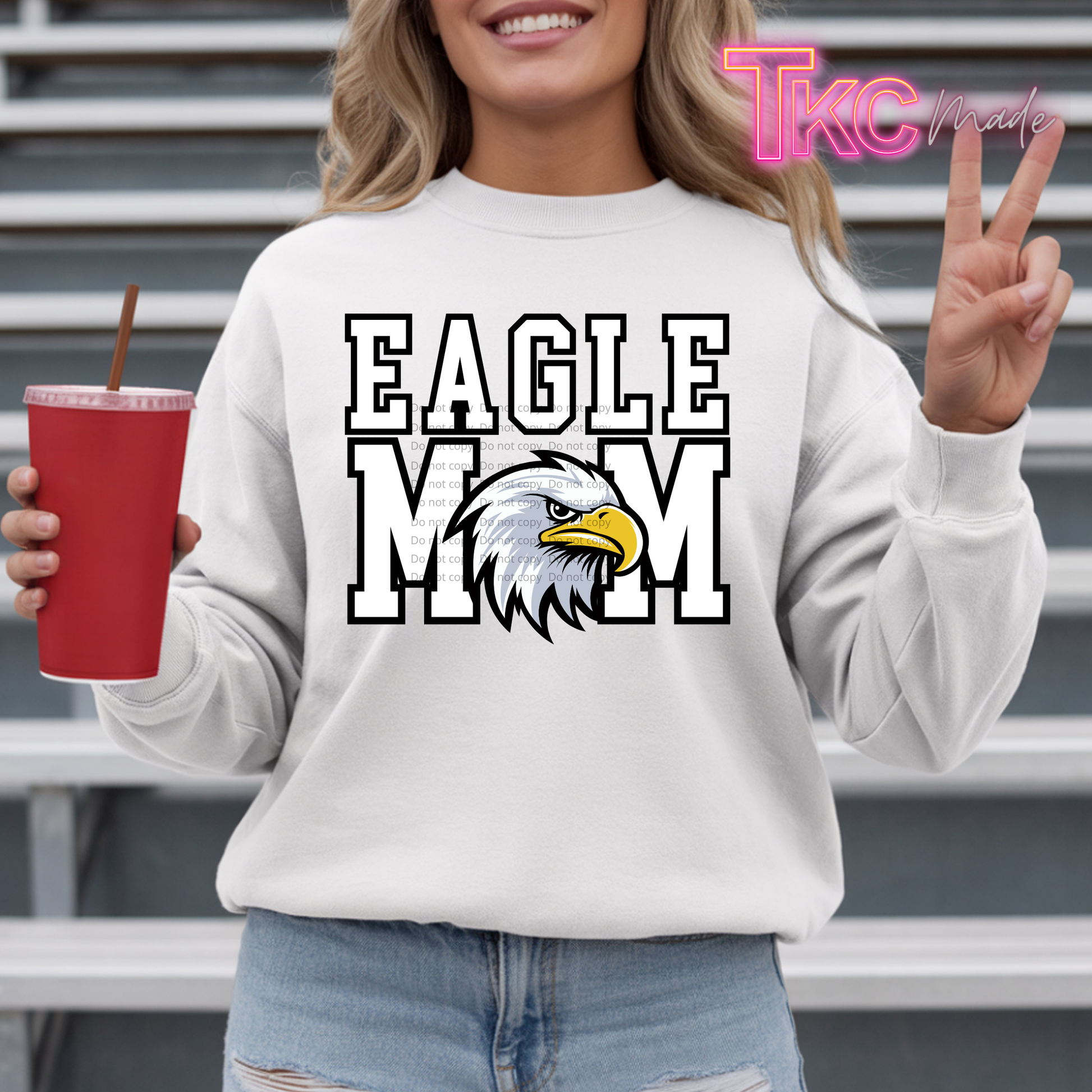 A woman wearing a white crewneck sweatshirt with a graphic that reads 'EAGLE MOM' in bold white letters with black outlines, featuring a bald eagle head design in the center. She is holding a red cup and making a peace sign, with metal bleachers in the background.