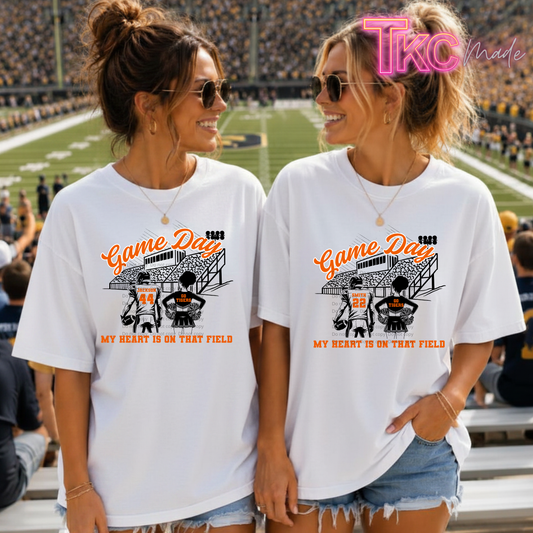 Two women standing together in a stadium wearing matching white graphic T-shirts with a football field illustration and the text 'Game Day ' and 'MY HEART IS ON THAT FIELD.' A pink 'TKC Made' watermark appears in the top right.