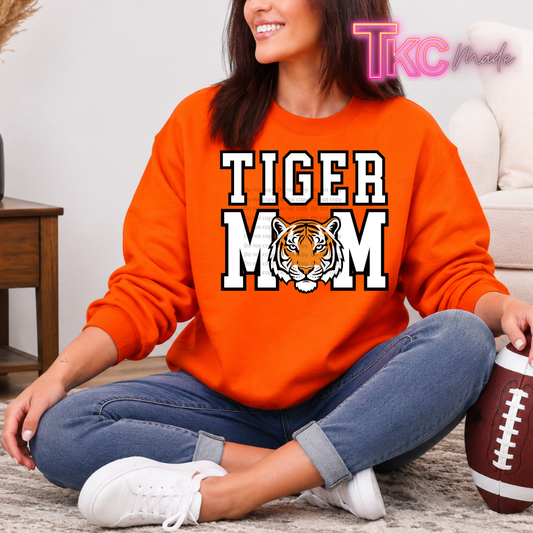 A woman sitting cross-legged on a rug wearing an orange crewneck sweatshirt with 'TIGER MOM' and a tiger graphic printed on the front. She is wearing blue jeans and white sneakers, with a football next to her in a cozy indoor setting.