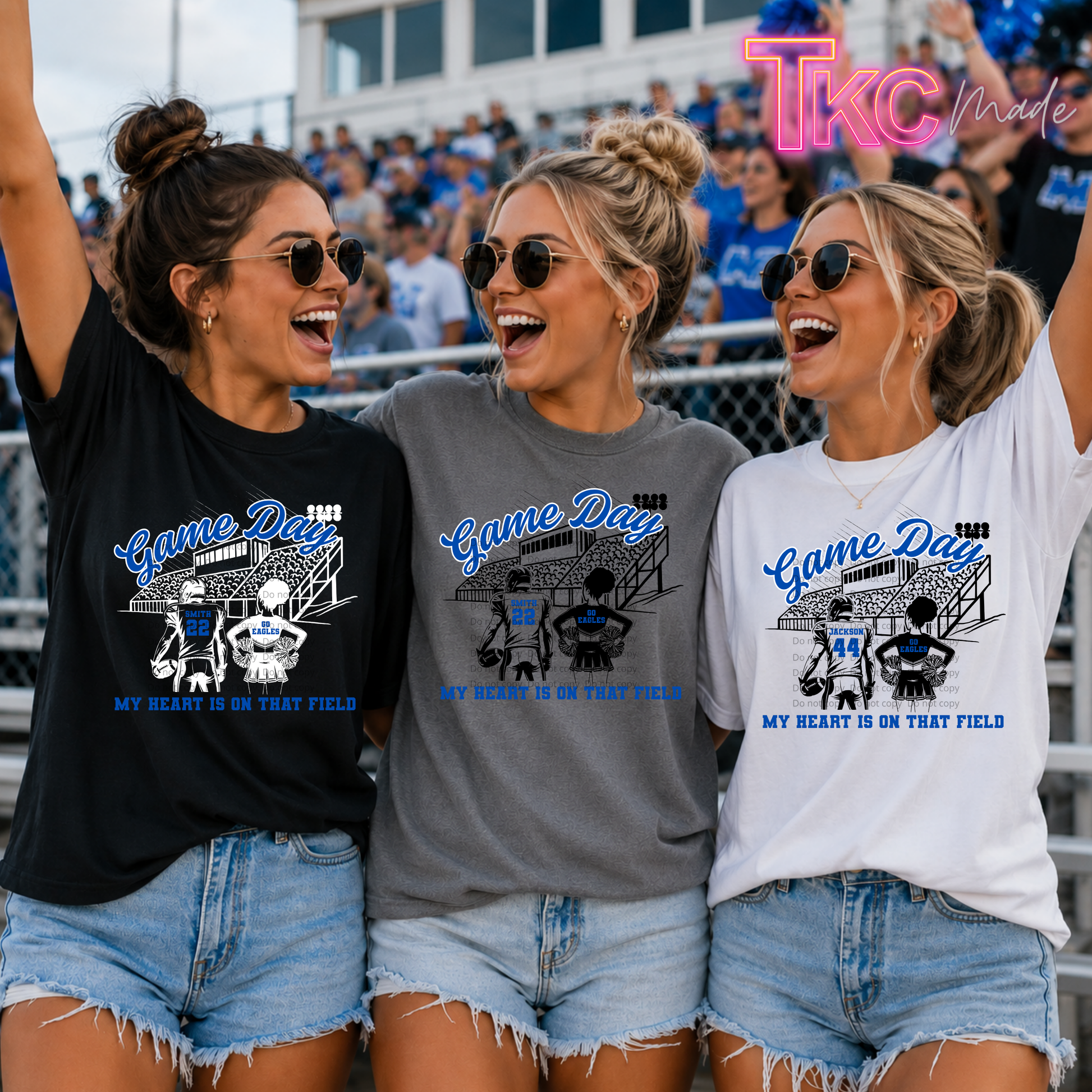 Three women wearing graphic t-shirts in black, gray, and white, standing together in stadium bleachers. The shirts feature a blue and white graphic of a football field and players with the text 'Game Day' and 'MY HEART IS ON THAT FIELD'.