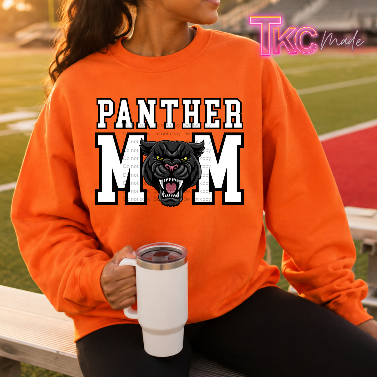 A woman wearing a bright orange sweatshirt with a 'PANTHER MOM' graphic featuring a black panther design, sitting on a bench at an outdoor sports field. She is holding a white travel mug, and a 'TKC Made' watermark is visible in the top right corner.