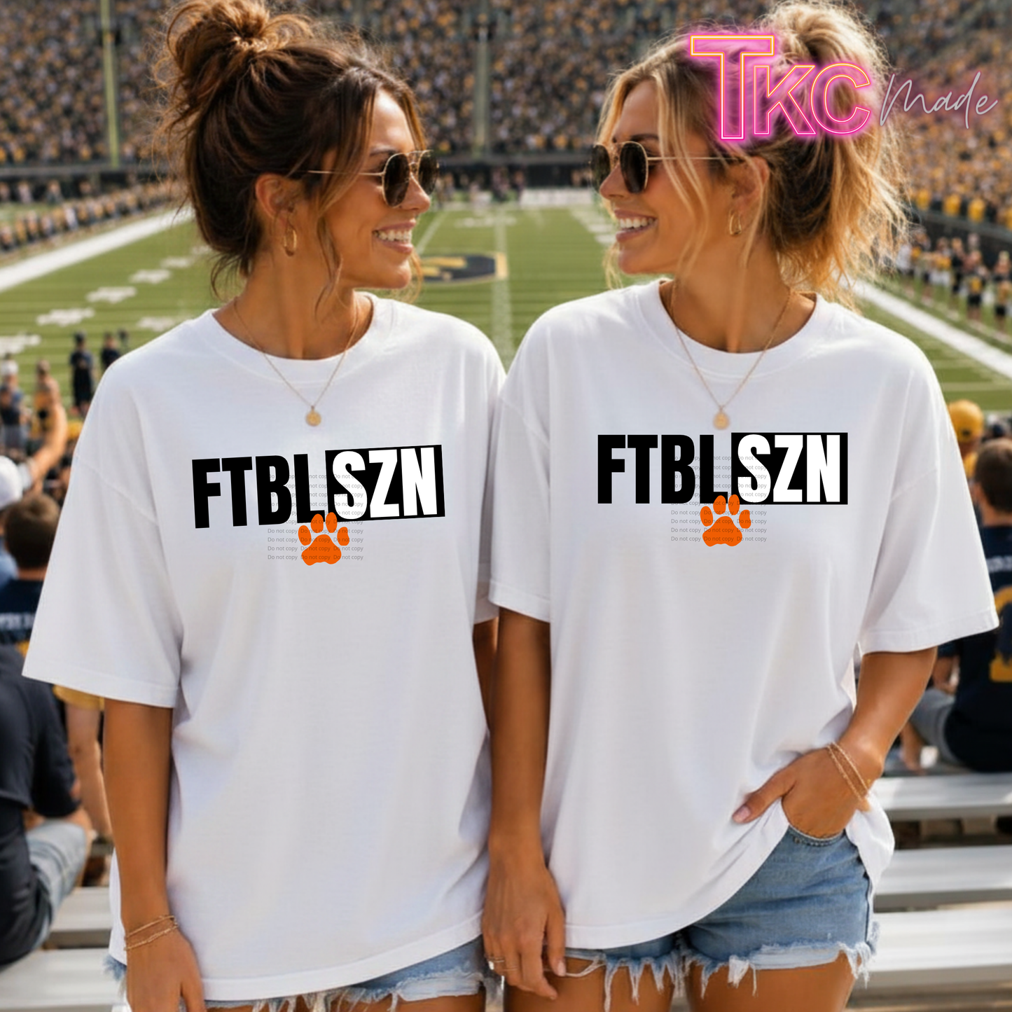 Two women standing in a stadium wearing matching white graphic T-shirts with the text 'FTBLSZN' in black and an orange paw print logo underneath. The background shows a football field and crowd.