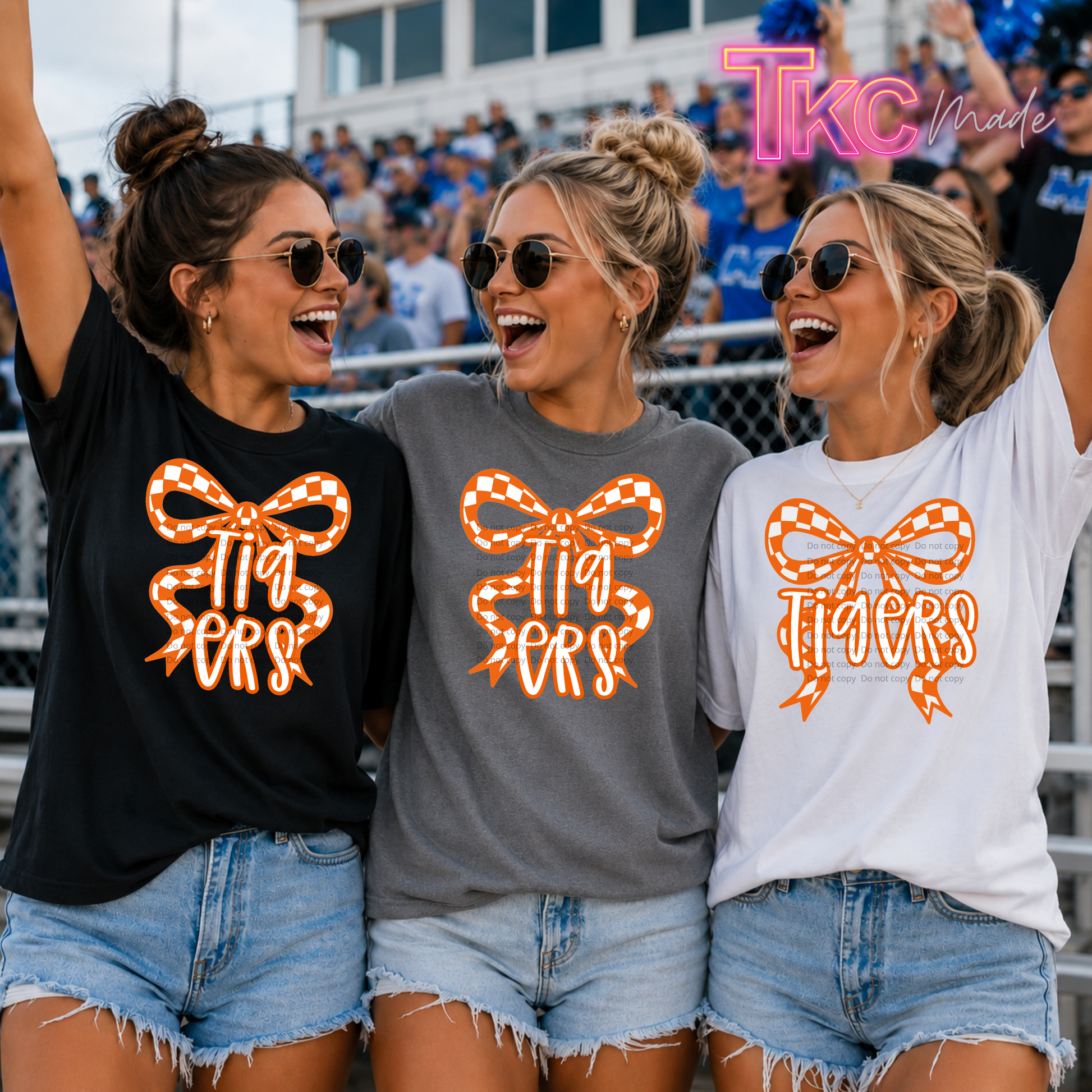 Three women wearing graphic t-shirts with a bow and 'Tigers' text design, standing together in a stadium crowd. The shirts are black, heather gray, and white, with the graphic in orange and white gingham.