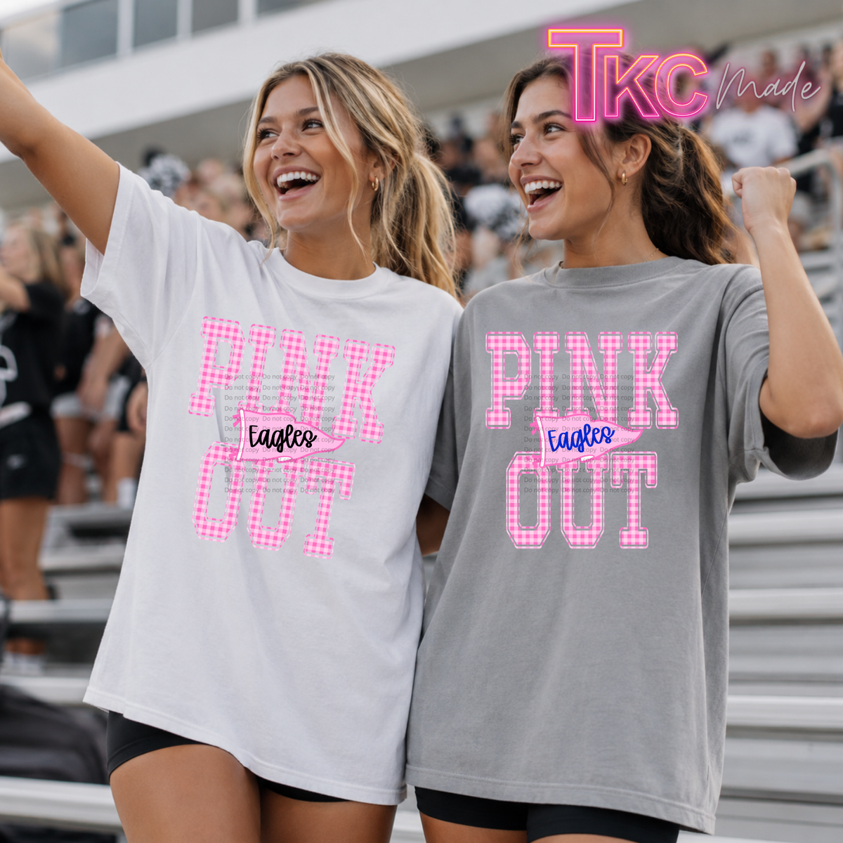 Two women standing in stadium seating wearing oversized graphic T-shirts with 'PINK OUT' in pink gingham letters and a pennant flag with the word eagles. One shirt is white and the other is heather gray.