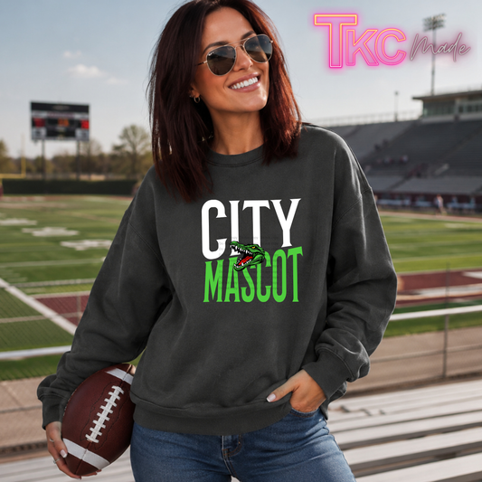 A woman wearing a dark charcoal gray sweatshirt with 'CITY MASCOT' printed in white and bright green letters, standing on a football field and holding a brown football. The sweatshirt features a small mascot graphic in the center of the text.