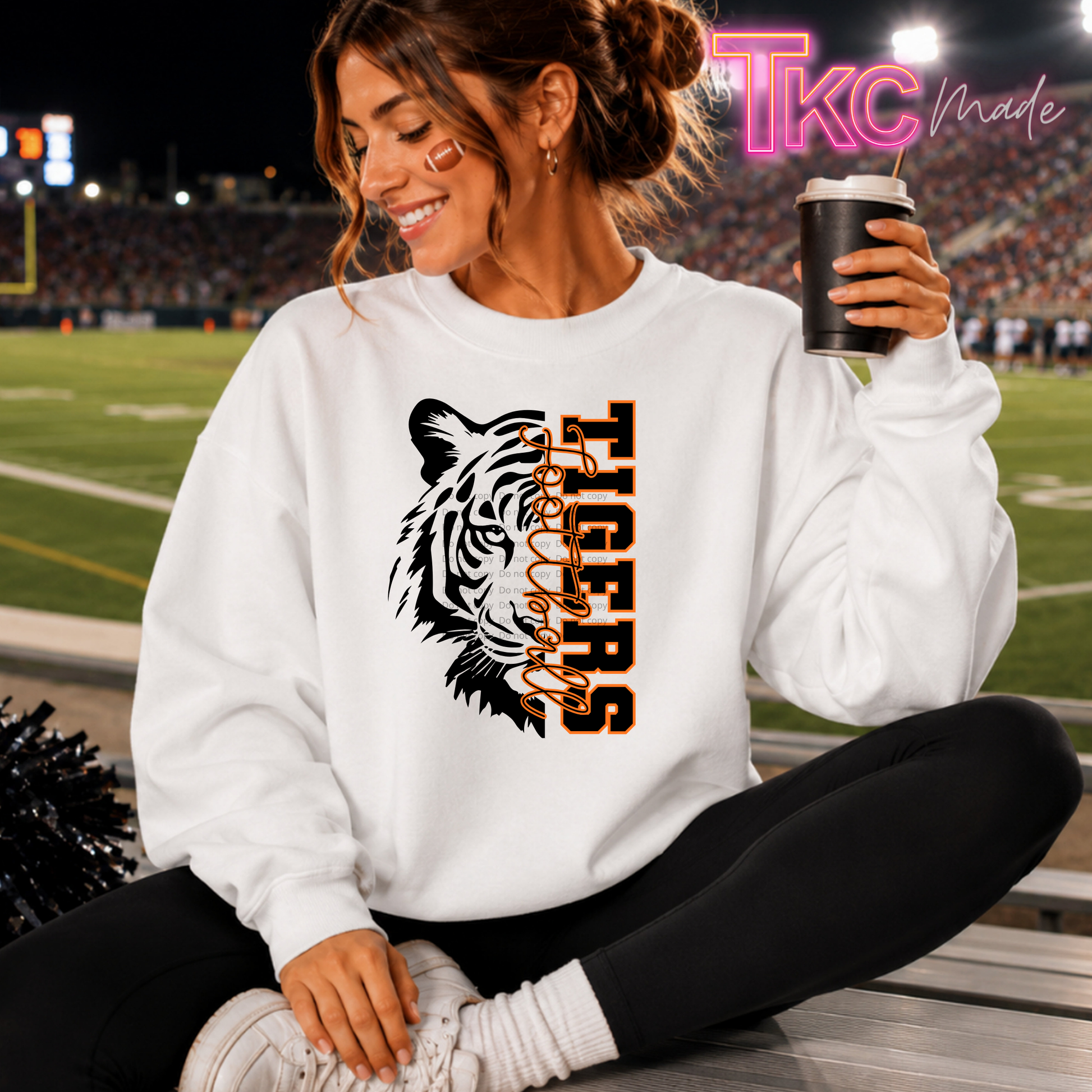 A woman sitting on a stadium bench at night wearing a white crewneck sweatshirt with a black and orange tiger graphic and the word 'TIGERS'. She is holding a black coffee cup, with a football field and stadium lights in the background.